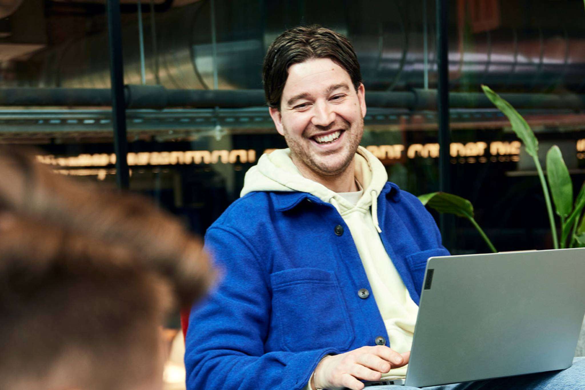 Man with laptop on lap looks smiling at a colleague.