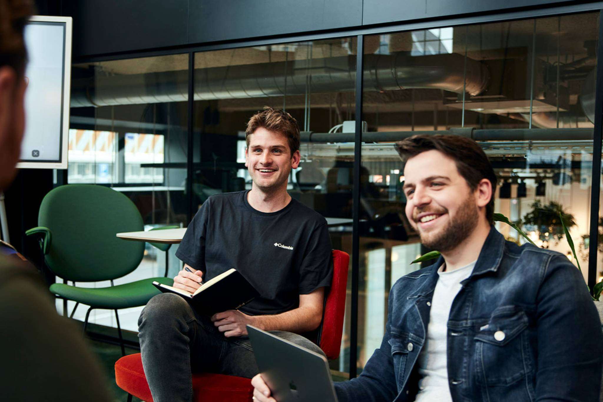 Man achter laptop lacht naar collega terwijl een andere man aantekeningen maakt.